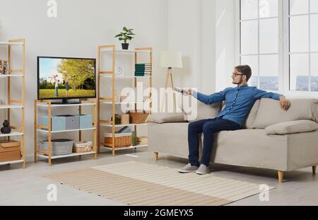 Giovane uomo che si rilassa sul divano in soggiorno e guarda i film sul grande schermo della TV moderna Foto Stock