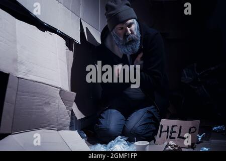 Un uomo senza tetto si siede sulle scatole per strada e chiede aiuto. Ha bisogno di una persona senza tetto chiede soldi per il cibo e durante la notte. Foto Stock