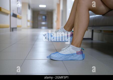 Donna in copriscarpe seduta in corridoio ospedale primo piano. Norme sanitarie di comportamento in istituzioni mediche concetto Foto Stock