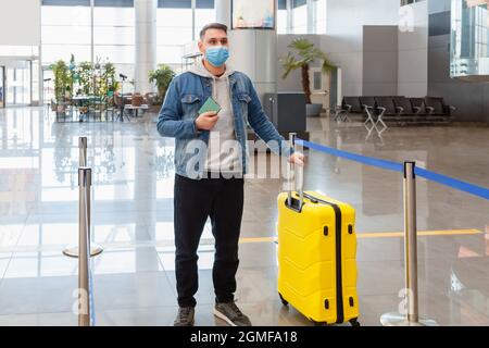 Uomo in maschera medica di fronte al banco check-in all'interno dell'aeroporto durante il blocco covid. Il viaggiatore informale urbano può trasportare il bagaglio della valigia del passaporto Foto Stock