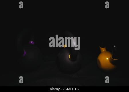 Tre kettlebells scuri su sfondo nero con punti di luce su di loro, due neri con marcature colorate, uno giallo Foto Stock
