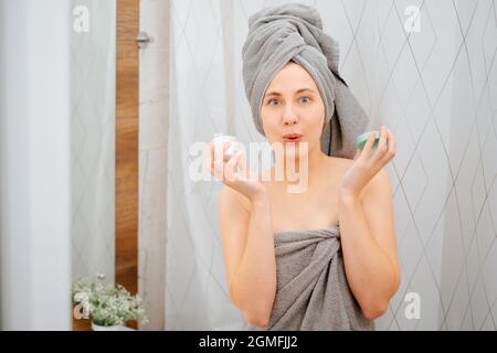 Donna in un asciugamano grigio sullo sfondo del bagno tiene un vaso di siero. Foto Stock