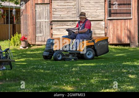 Velena, Lettonia - 19 giugno 2021: Un uomo anziano con un rasaerba rasa l'erba nel cortile della casa di campagna Foto Stock