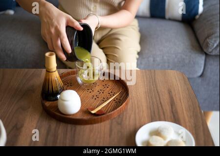 Donna che versa pronto matcha giapponese verde tè bevanda a casa utilizzando tradizionale tè cerimonia bambù set mentre seduto sul divano a casa. Foto Stock