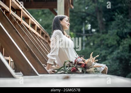 Una giovane donna in abito bianco si siede su un ponte di legno con un bouquet di fiori esotici. Foto Stock