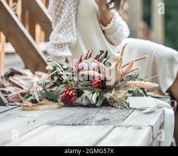 Una giovane donna in abito bianco siede su un ponte di legno con un bouquet di esotici fiori protea. Foto Stock