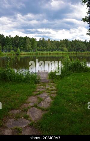 Un sentiero in pietra conduce ad un piccolo molo di legno sulle rive di un pittoresco fiume. Sullo sfondo la foresta estiva e le nuvole di pioggia Foto Stock