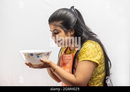 Una donna casalinga indiana graziosa in grembiule di cottura che guarda una ciotola di servizio in mano su sfondo bianco Foto Stock