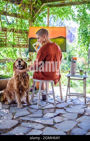 Giovane artista femminile che lavora sulla sua tela d'arte dipingendo all'aperto nel suo giardino con Golden Retriever mantenendo la sua azienda. Concetto creativo di hobby. Foto Stock