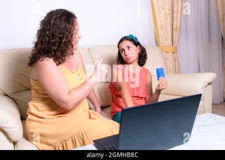 Bruna ragazza seduta sul divano a casa, facendo un volto pietoso per comprare qualcosa per lei online, ma la madre punta il dito contro di lei e dice no Foto Stock