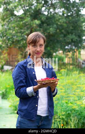 Una bella giovane donna in una camicia blu tiene in mano un cesto di lamponi freschi e profumati su uno sfondo di verde fogliame. Foc. Selettivo Foto Stock