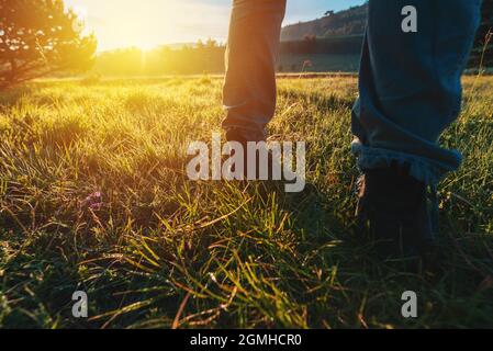 Escursioni nella prateria, primo piano di piedi maschi in stivali di pelle che cammina attraverso il paesaggio erboso Zlatibor all'alba, fuoco selettivo. Foto Stock