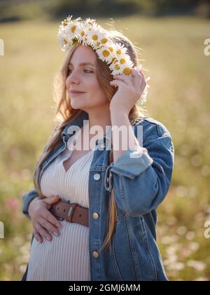 Bella donna incinta in un abito, giacca in denim e una corona di margherite rilassante fuori nel parco. Gravidanza sana e concetto di viaggio. Foto Stock