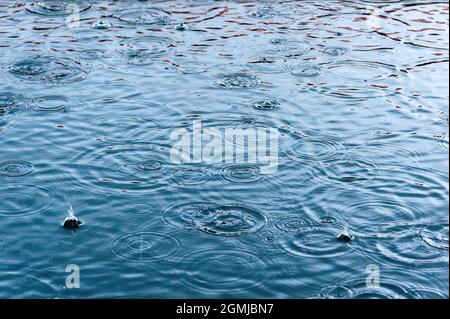 la pioggia cade sulla superficie dell'acqua in una pozza con sfumatura graduata di ombra nera e riflesso del cielo blu Foto Stock