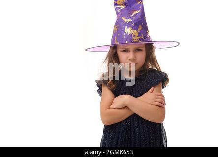 Ritratto di bella piccola strega ragazza arrabbiata con un cappello mago e vestito in elegante abito carnevale, guardando la macchina fotografica in posa con braccio incrociato Foto Stock