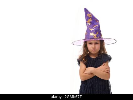 Ritratto di winsome piccola strega arrabbiata ragazza sconvolta indossando un cappello mago e vestito in elegante abito carnevale, guardando la macchina fotografica in posa con l'ar incrociato Foto Stock