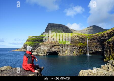 Un turista in giacca rossa guarda una cascata Mulafossur nel villaggio di Gasadalur, Isole Faroe Foto Stock