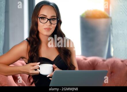 Una bella giovane donna europea in bicchieri di 20-30 anni si siede sul divano e tiene una tazza di caffè o tè. Una bella ragazza lavora in remoto su un laptop. Foto Stock