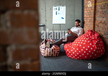 Giovane uomo d'affari con smartphone e cuffie che fa una pausa in ufficio, riposando. Foto Stock