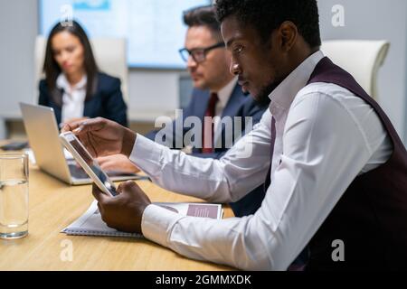 Un uomo d'affari serio che scorre in un tablet mentre è seduto al tavolo contro i suoi colleghi Foto Stock