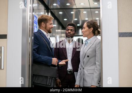 Giovane imprenditore africano che guarda i suoi colleghi interagenti in ascensore Foto Stock