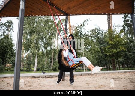 Allegra madre musulmana in hijab in piedi vicino alla figlia in altalena nel parco Foto Stock