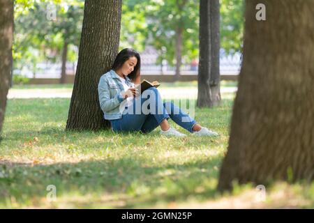 Concetto di detox digitale - donna asiatica che legge un libro nel parco. Si siede sull'erba sotto l'albero, legge un libro e prende una pausa dalla costantl Foto Stock