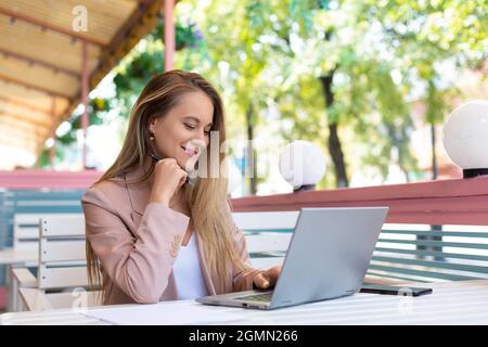 Donna che lavora davanti al monitor del portatile al bar estivo. Guarda il monitor del notebook e sorride. Tecnologia moderna nel concetto di vita di ogni giorno. Foto Stock