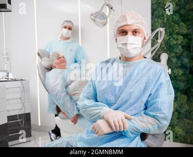 Ritratto del dentista dell'uomo e del suo collega in piedi accanto alla paziente femminile. Donna in cappuccio medico monouso giacente in sedia dentale mentre ha appuntamento con i dentisti in clinica dentale moderna. Foto Stock