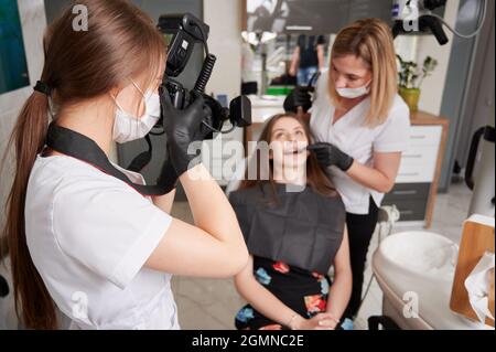 Dentista che esamina i denti della donna mentre assistente nella maschera medica che scatta la foto con la macchina fotografica professionale. Giovane medico femminile in guanti sterili fotografando paziente e dentista in studio dentistico. Foto Stock