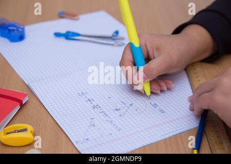 La mano della pupilla del primo piano scrive una formula geometrica in un taccuino. Uno scolaro svolge un compito sul posto di lavoro. Il concetto di educazione dei bambini, di insegnamento delle conoscenze, delle competenze e delle abilità. Foto Stock