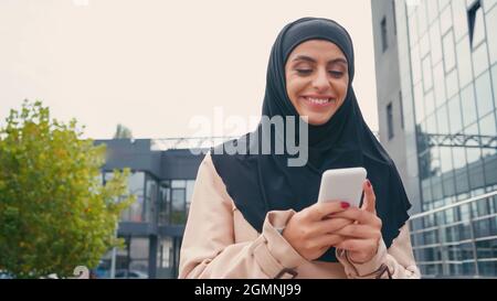 ragazza musulmana allegra in messaggi hijab sul telefono cellulare esterno Foto Stock