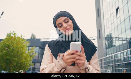 donna musulmana sorridente in messaggi hijab su smartphone all'esterno Foto Stock