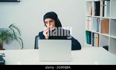 Donna d'affari musulmana in cuffia utilizzando il microfono vicino al laptop nel call center Foto Stock