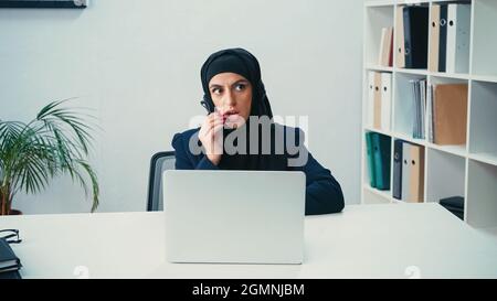 Donna musulmana in cuffia utilizzando il microfono vicino al laptop nel call center Foto Stock