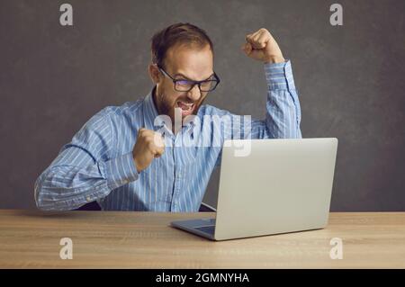 Un uomo d'affari entusiasta seduto al tavolo con un computer portatile, che tira il pugno e gridava Sì Foto Stock