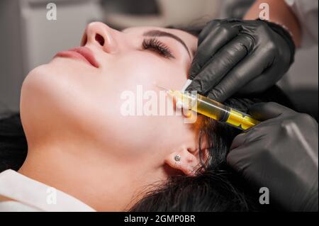 Primo piano della procedura di iniezione del plasmolifting. Medico cosmetologo estetista in guanti neri sterili facendo l'iniezione in volto femminile paziente. Concetto di trattamento della pelle. Foto Stock