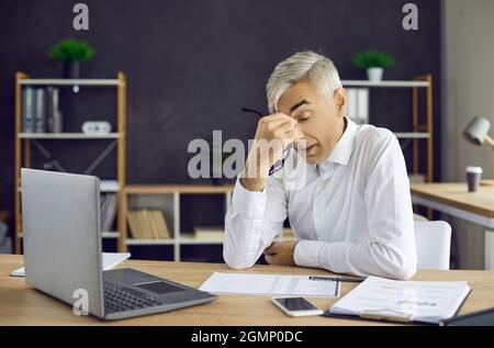 L'uomo stanco e stressato seduto alla scrivania dell'ufficio con un computer portatile toglie gli occhiali e gli occhi di sfregio Foto Stock