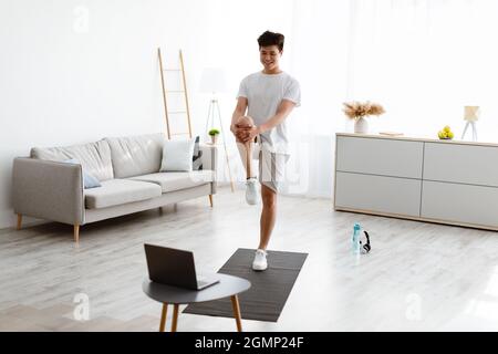 Ragazzo asiatico che fa in piedi ginocchio al petto esercizio fisico utilizzando il portatile Foto Stock