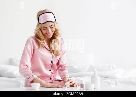 Felice cute donna bionda caucasica in pajama rosa con maschera di sonno messo su smalto unghie Foto Stock