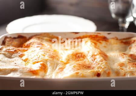 Lasagne fatte in casa in un recipiente bianco con piatti e bicchieri sullo sfondo, primo piano Foto Stock