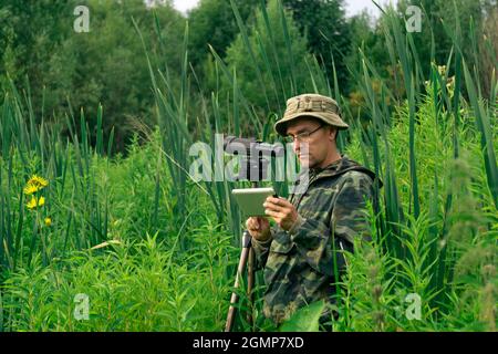 l'uomo birdwatcher guarda o scrive le informazioni sulla tavoletta mentre si trova in piedi tra l'erba alta nella zona umida Foto Stock