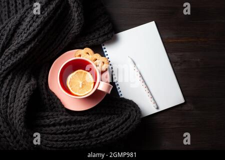 Copybook trasparente, tazza di tè melograno, penna e sciarpa a maglia su sfondo di legno nero Foto Stock