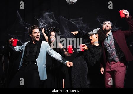 donna afroamericana in costume di halloween che punta con le dita mentre balla con eccitati amici multietnici su nero Foto Stock