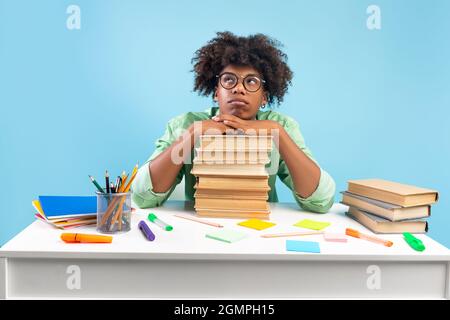 Triste studente africano americano maschile seduto a tavola e appoggiato su una pila di libri, non vuole studiare, sfondo blu Foto Stock