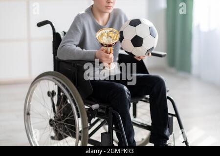 Vista ritagliata del giocatore di calcio teen in sedia a rotelle che tiene palla e trofeo, sentendosi stressato sopra il suo infortunio a casa Foto Stock