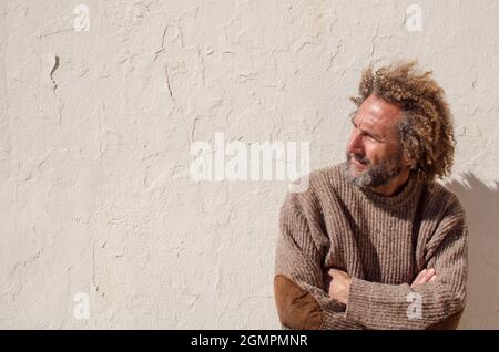 uomo anziano di 50-60 anni con capelli lunghi appoggiati a una parete bianca guarda al lato, all'orizzonte con le braccia incrociate Foto Stock