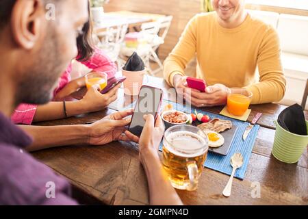Primo piano di un uomo irriconoscibile utilizzando il suo cellulare in un ristorante, caffè. Gruppo di amici che si divertono a condividere online. Foto Stock