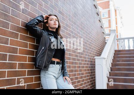 giovane donna sognante in giacca e jeans in pelle accanto a muro di mattoni Foto Stock