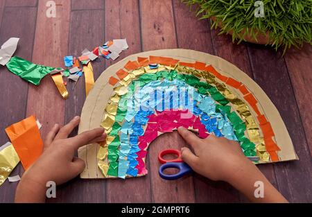 le mani del bambino fanno un arcobaleno con carta colorata e una forbice su un tavolo di legno per celebrare la primavera. orizzontale. Attività per bambini a casa Foto Stock
